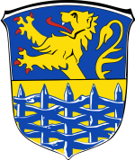 In diesem Bild sehen Sie das Wappen der Samtgemeinde Hage. Der Hintergrund ist hellblau und gelb. Im Vordergrund ist ein hellblauer Zaun zu sehen. Hinter dem Zaun befindet sich ein gelber Löwe. 