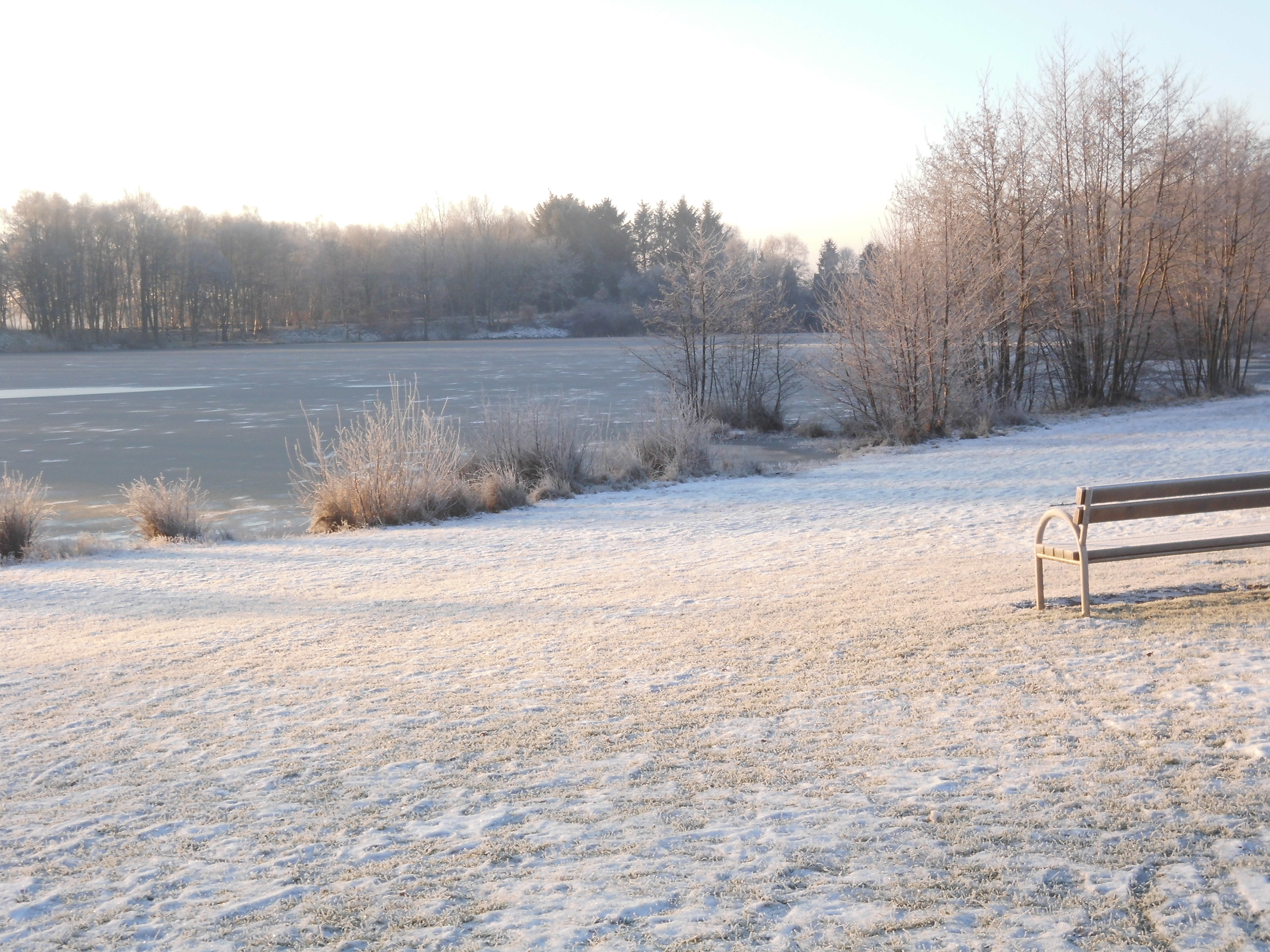 Auf dem Foto ist im Hintergrund der Kiessee von Berum im Winter zu erkennen. Es liegt leichter Schnee. Auf der rechten Seite befindet sich eine Parkbank. Die Sonne scheint aufzugehen.