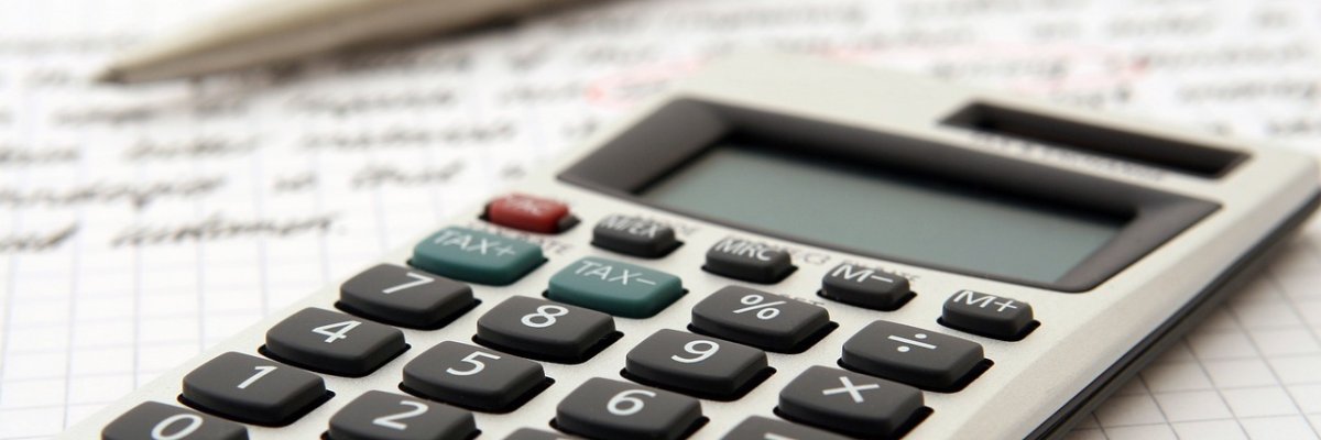 Grauer Taschenrechner vorne im Bild auf einem beschriebenen Blatt Papier. Im Hintergrund liegt ein silberner Kugelschreiber.